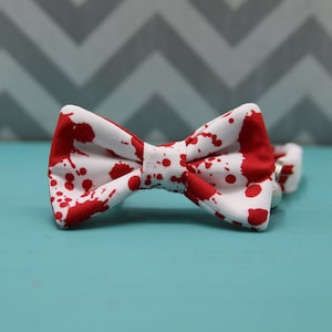 May include: A white bow tie with a red splatter pattern. The bow tie is made of fabric and has a classic bow shape. The background is a light blue surface with a gray and white chevron pattern.
