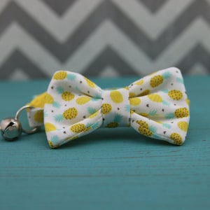 May include: A white bow tie with a yellow pineapple pattern and a bell on a yellow strap. The bow tie is on a teal blue wooden surface.