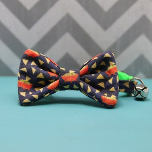 May include: A blue cat collar with a bow tie featuring a pattern of red and yellow tortilla chips and green guacamole. The bow tie is attached to a green collar with a silver bell.