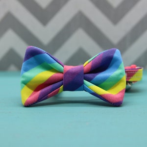 May include: A colorful rainbow-striped bow tie for pets. The bow tie features vibrant bands of red, orange, yellow, green, blue, and purple. The bow tie is attached to a matching collar with a pink and yellow pattern.