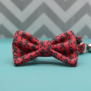 May include: A red fabric bow tie featuring a repeating pattern of small black stick figures in various poses. The bow tie has a central knot and is attached to a matching fabric strap with a metal buckle and a small metal ring on the side.