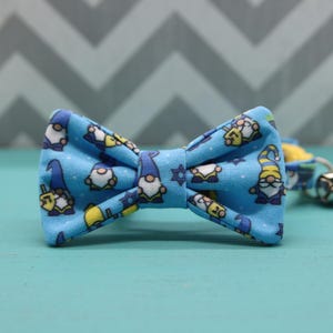 May include: A blue bow tie with a pattern of gnomes wearing yarmulkes and holding dreidels. The bow tie is made of fabric and has a star of David in the center.