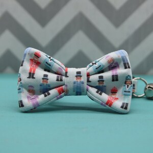 May include: A light blue fabric bow tie featuring a repeating pattern of small nutcracker figures in various colors including red, blue, and purple. The bow tie has a central fabric knot and a small metal ring with a bell attached to the side.