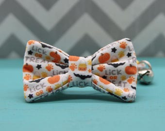 May include: A white bow tie with a Halloween-themed print featuring orange pumpkins, black bats, and the text "Lick or Treat". The bow tie has a bell attached.
