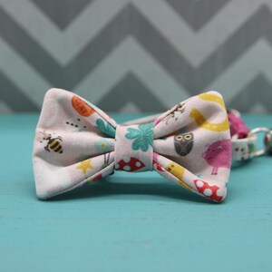 May include: A light pink bow tie with a whimsical pattern of bees, mushrooms, owls, and flowers. The bow tie is made of fabric and has a matching collar. The background is a blue surface with a gray chevron pattern.