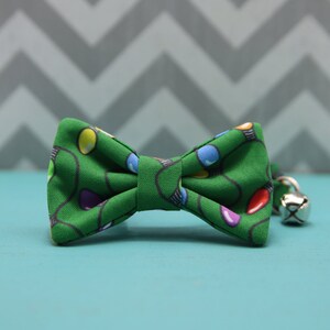 May include: A green bow tie with a pattern of colorful Christmas lights. The bow tie has a bell attached to it.