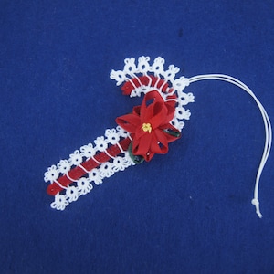 Puede incluir: Adorno de bastón de caramelo de crochet blanco con un bastón de caramelo de fieltro rojo y una flor de poinsettia de fieltro rojo.