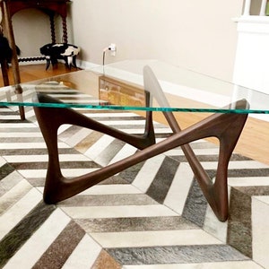 May include: A triangular glass top coffee table with a unique, mid-century modern, dark wood base. The table is sitting on a black and white chevron patterned rug.