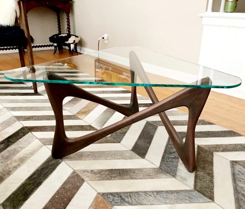 May include: A triangular glass top coffee table with a unique, mid-century modern, dark wood base. The table is sitting on a black and white chevron patterned rug.