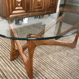 May include: A mid-century modern glass top coffee table with a unique, organic wood base. The table has a clear glass top and a natural wood finish.