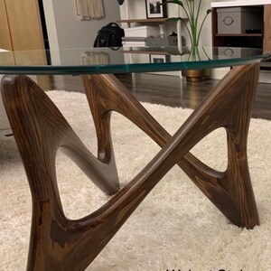 May include: A glass-top coffee table with a unique, abstract, wooden base. The base is stained a dark walnut color and is made of fir wood.
