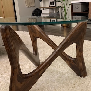 May include: A close-up of a glass-top coffee table with a unique, abstract wooden base. The base is stained a dark walnut color and has a natural wood grain pattern. The text "Walnut Stain on Fir Wood" is visible below the table.
