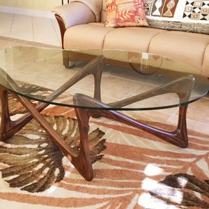 May include: A mid-century modern glass and wood coffee table with a unique, sculptural base. The table has an oval glass top and a dark wood base with a geometric design. The table is sitting on a patterned rug.