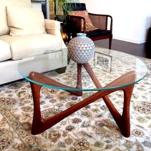 May include: A glass-top coffee table with a unique, X-shaped wooden base. The table has a light wood finish and sits on a patterned rug. A blue and white ceramic vase sits on the table.