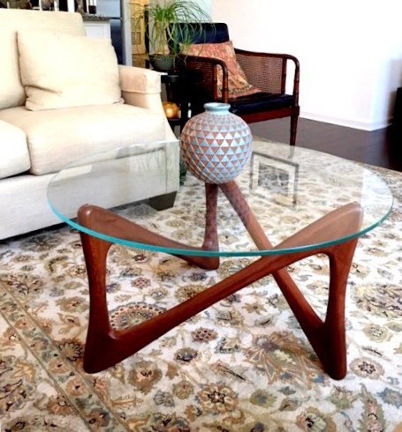 May include: A glass-top coffee table with a unique, X-shaped wooden base. The table has a light wood finish and sits on a patterned rug. A blue and white ceramic vase sits on the table.
