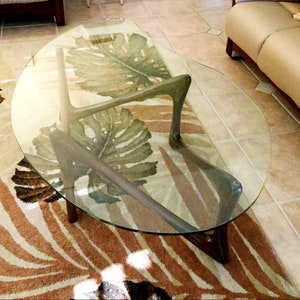 May include: A glass-top coffee table with a unique, curved, wooden base. The table is sitting on a rug with a leafy pattern.