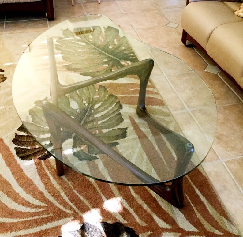 May include: A glass-top coffee table with a unique, curved, wooden base. The table is sitting on a rug with a leafy pattern.