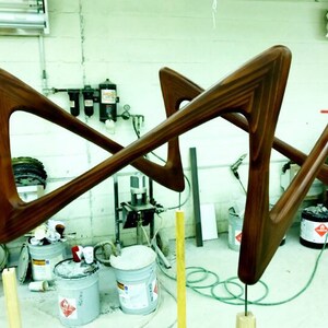May include: Two wooden frames in a workshop setting. The frames are made of dark brown wood and have a unique, geometric shape. The frames are suspended from the ceiling by wires and are being prepared for finishing.