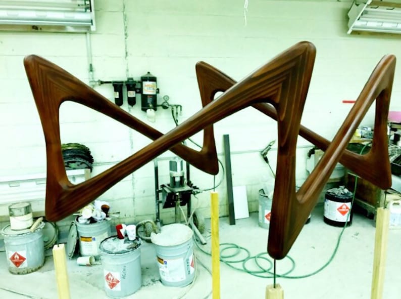 May include: Two wooden frames in a workshop setting. The frames are made of dark brown wood and have a unique, geometric shape. The frames are suspended from the ceiling by wires and are being prepared for finishing.