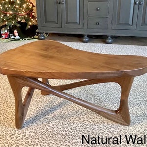 May include: A natural walnut wood coffee table with a unique, asymmetrical design. The table has a wide, flat top and three legs that curve and intersect in a modern style.