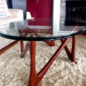May include: A round glass coffee table with a dark wood base. The base has a unique, abstract design with three legs. The table is sitting on a white shag rug.