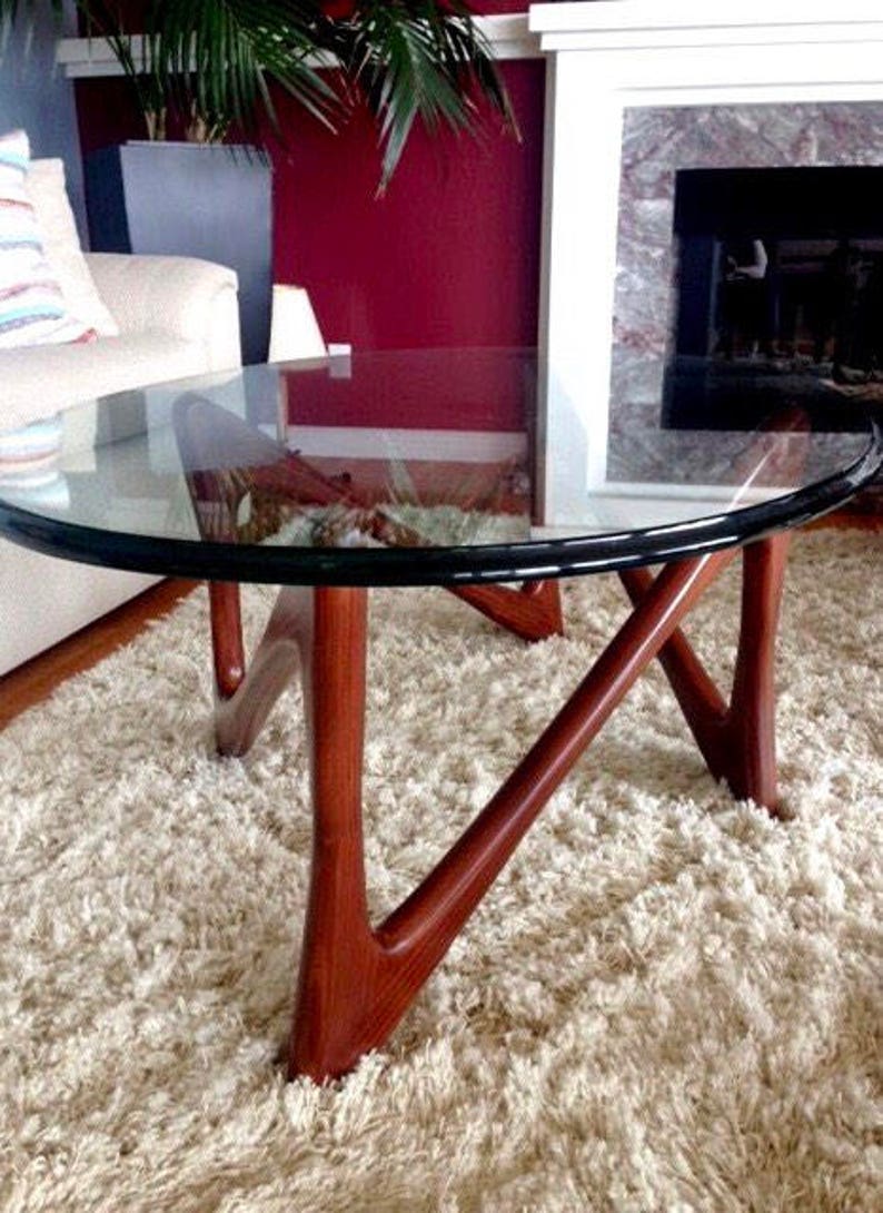 May include: A round glass coffee table with a dark wood base. The base has a unique, abstract design with three legs. The table is sitting on a white shag rug.