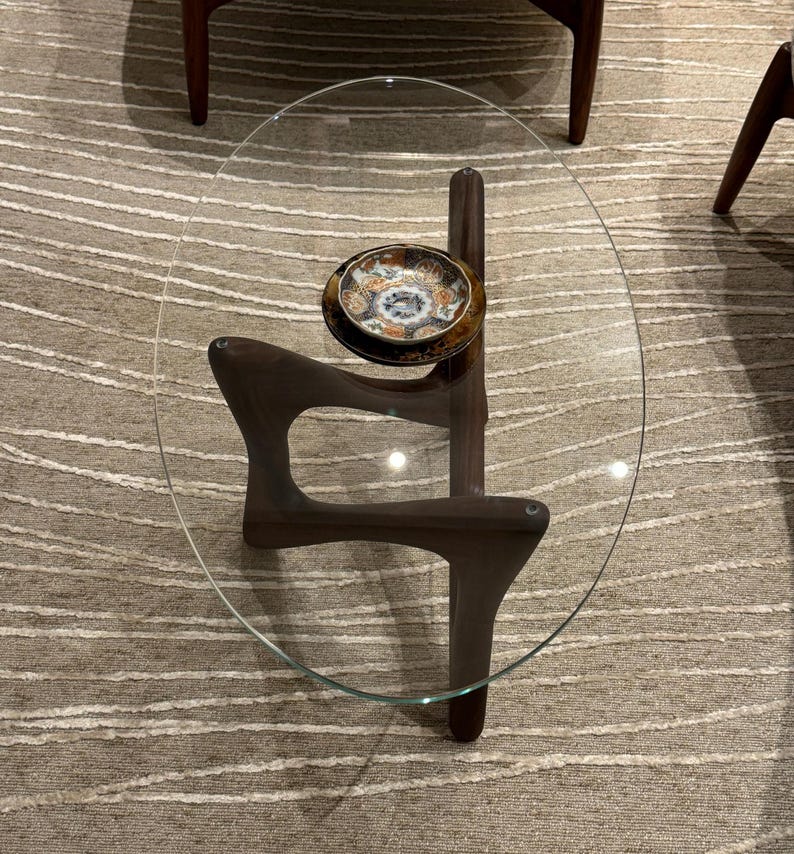 Pu&ograve; includere: Un tavolino da caff&egrave; ovale in vetro con una base in legno scuro e un piatto decorativo in cima.