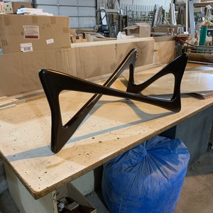 May include: A dark brown wooden table base with a unique X-shaped design. The base is sitting on a light brown wooden surface. A blue bag is in the foreground.