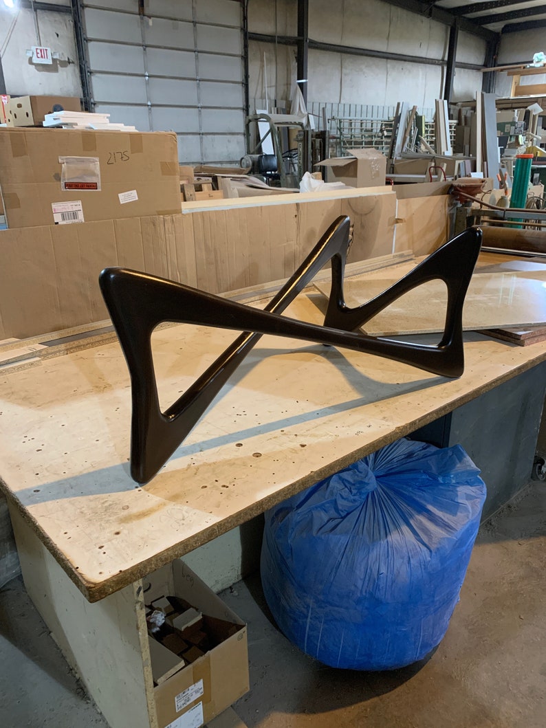 May include: A dark brown wooden table base with a unique X-shaped design. The base is sitting on a light brown wooden surface. A blue bag is in the foreground.