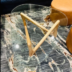 May include: A glass-top coffee table with a triangular wooden base. The table has a curved, oval-shaped top and sits on a patterned rug.