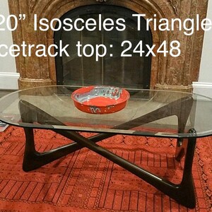 May include: A glass-top coffee table with a dark wood base. The base is shaped like an isosceles triangle with a racetrack top. The table is 34.5 inches by 20 inches and the top is 24 inches by 48 inches.