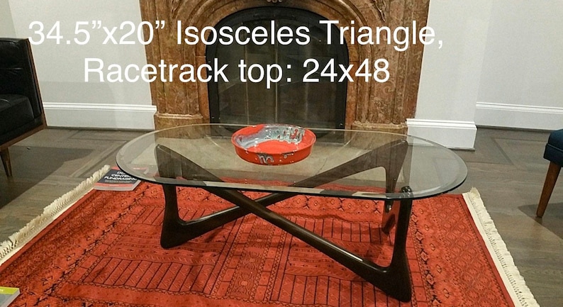 May include: A glass-top coffee table with a dark wood base. The base is shaped like an isosceles triangle with a racetrack top. The table is 34.5 inches by 20 inches and the top is 24 inches by 48 inches.