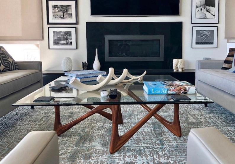 May include: A glass coffee table with a unique, modern design. The table has a clear glass top and a wooden base with a geometric shape. The table is sitting on a gray and white rug in a living room setting.