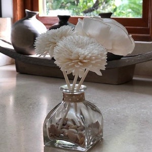 May include: A clear glass diffuser bottle with white wooden sticks and white paper flowers. The bottle is filled with white stones and the flowers are arranged in a bouquet.