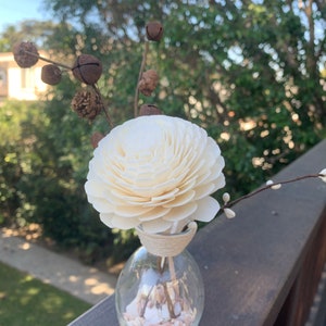 May include: A white wooden flower diffuser with a brown twig and small bells, in a clear glass bottle filled with white and pink seashells.