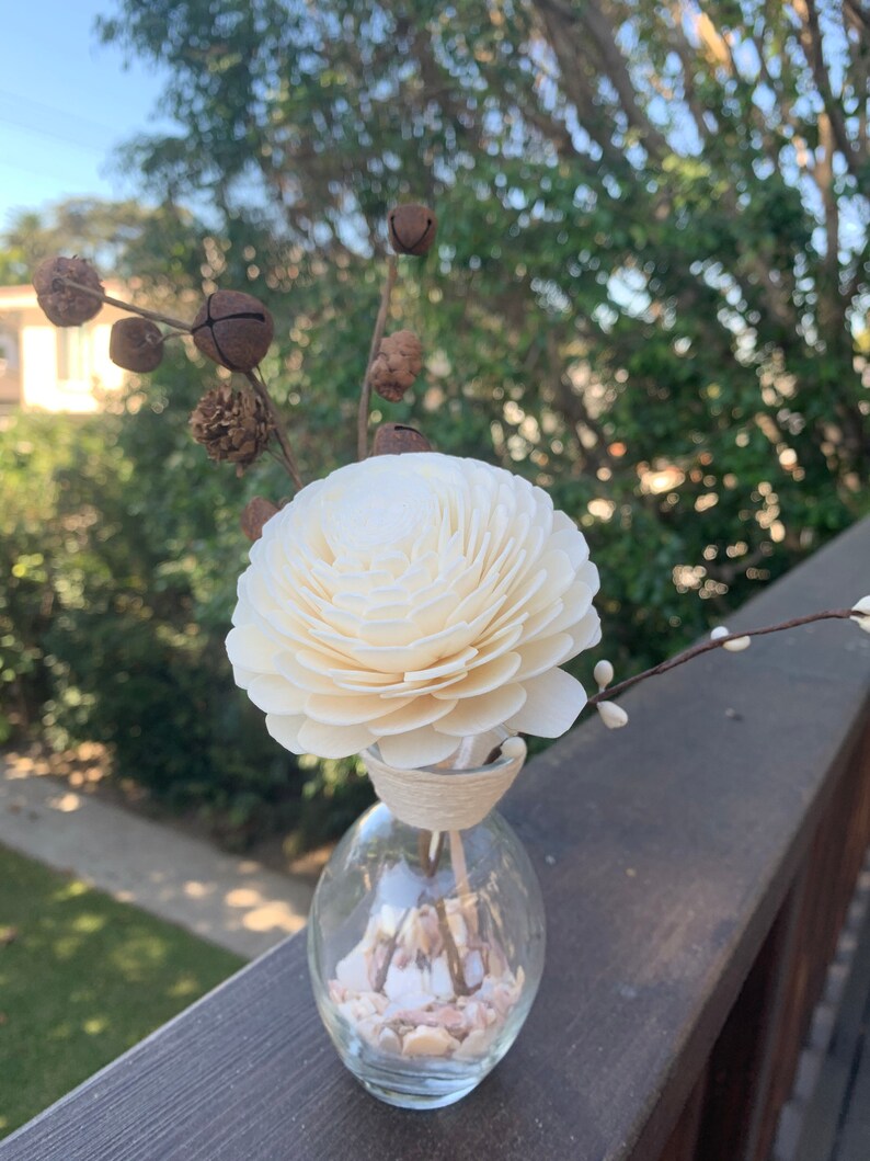May include: A white wooden flower diffuser with a brown twig and small bells, in a clear glass bottle filled with white and pink seashells.