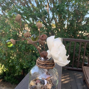May include: A clear glass vase with a rustic brown twine wrap around the neck. The vase is filled with small, smooth stones and a single white flower. The vase is decorated with a sprig of faux greenery with green acorn-shaped ornaments.