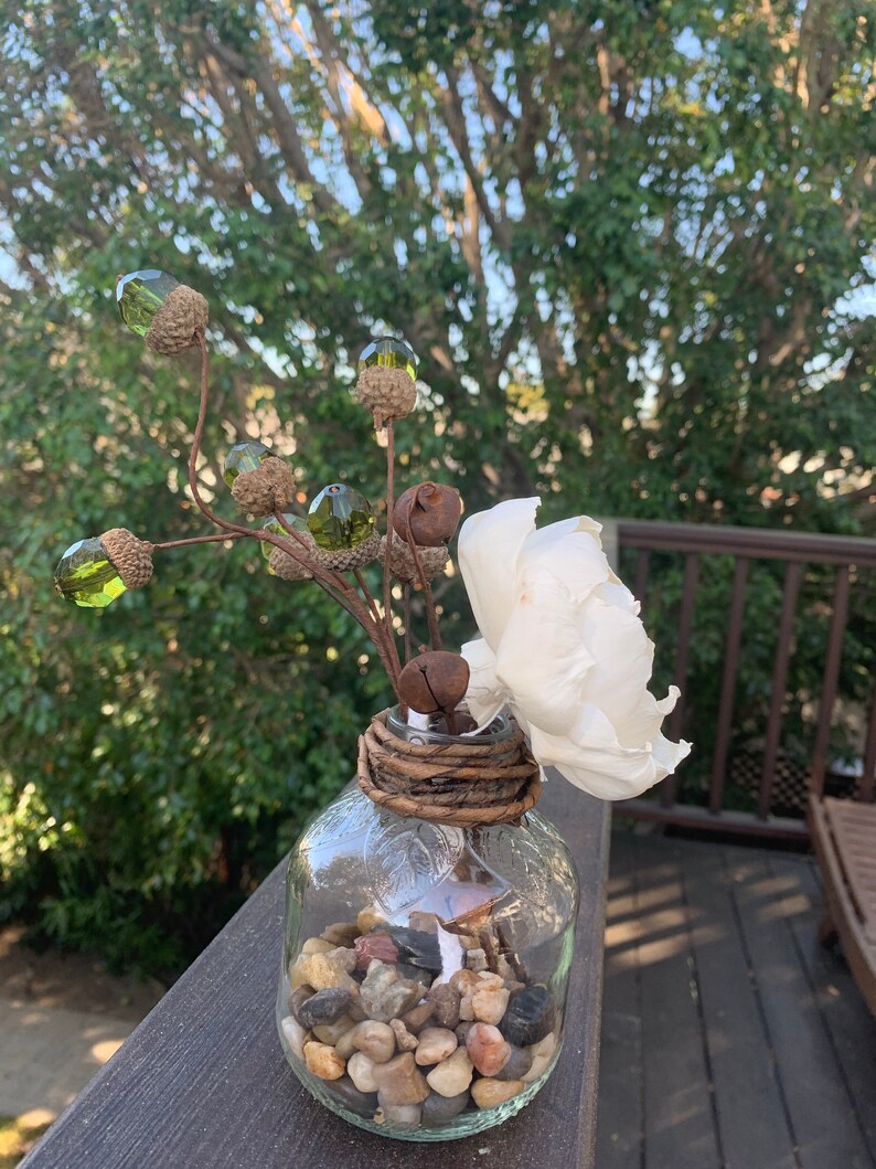 May include: A clear glass vase with a rustic brown twine wrap around the neck. The vase is filled with small, smooth stones and a single white flower. The vase is decorated with a sprig of faux greenery with green acorn-shaped ornaments.