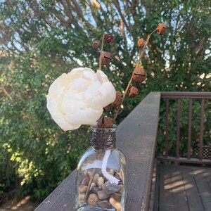 May include: A white faux flower arrangement in a clear glass vase with small stones and a white wick. The vase is decorated with brown twine and rusty bells.