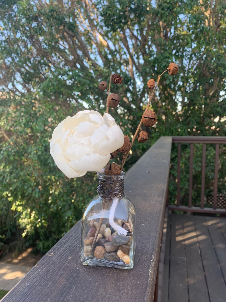 May include: A white faux flower arrangement in a clear glass vase with small stones and a white wick. The vase is decorated with brown twine and rusty bells.