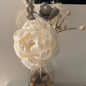 May include: A white faux flower arrangement in a clear glass vase with brown stones. The arrangement includes a large white flower, a grey flower, and white berry sprigs.