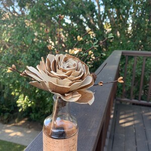 May include: A wooden rose in a clear glass bottle with a cork label. The bottle is sitting on a wooden surface.