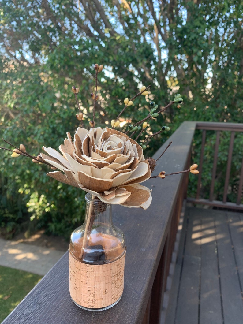 May include: A wooden rose in a clear glass bottle with a cork label. The bottle is sitting on a wooden surface.