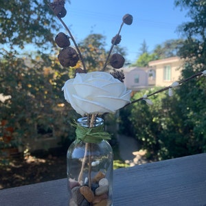 May include: A white faux rose with dried branches and brown seed pods in a clear glass bottle filled with small stones.