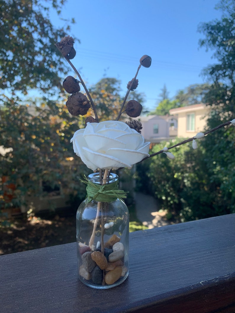 May include: A white faux rose with dried branches and brown seed pods in a clear glass bottle filled with small stones.