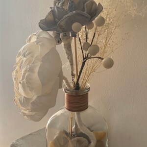 May include: A clear glass vase with a brown rope around the neck, filled with white and grey dried flowers and white decorative balls. The vase is sitting on a white textured surface.