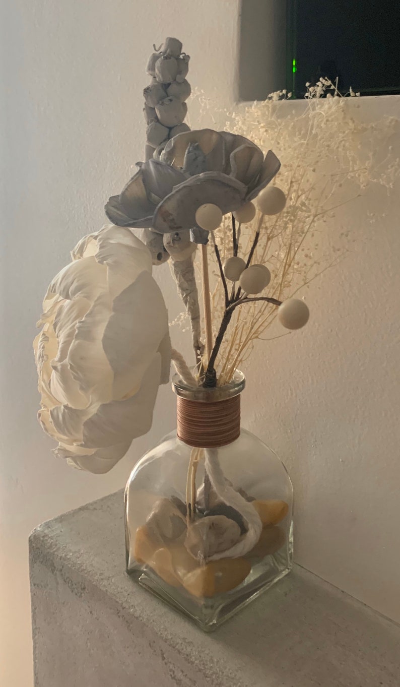 May include: A clear glass vase with a brown rope around the neck, filled with white and grey dried flowers and white decorative balls. The vase is sitting on a white textured surface.