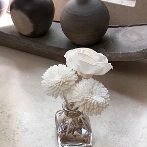 May include: A clear glass vase with a square base filled with small white stones and topped with three white dried flower arrangements. The vase is sitting on a white surface.