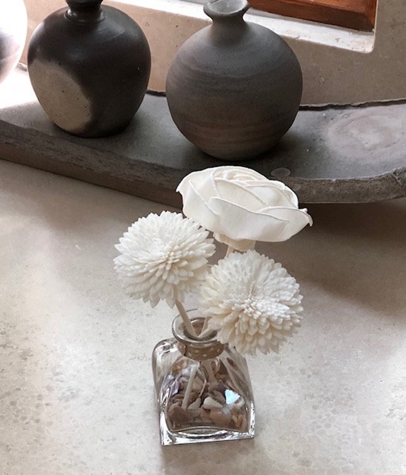 May include: A clear glass vase with a square base filled with small white stones and topped with three white dried flower arrangements. The vase is sitting on a white surface.