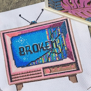 Puede incluir: Punto de cruz de un televisor retro con un marco rosa y la palabra "BROKEN" en la pantalla. La pantalla muestra un efecto pixelado y con fallos en azul, amarillo y verde. El televisor tiene dos antenas y está sobre tela blanca.